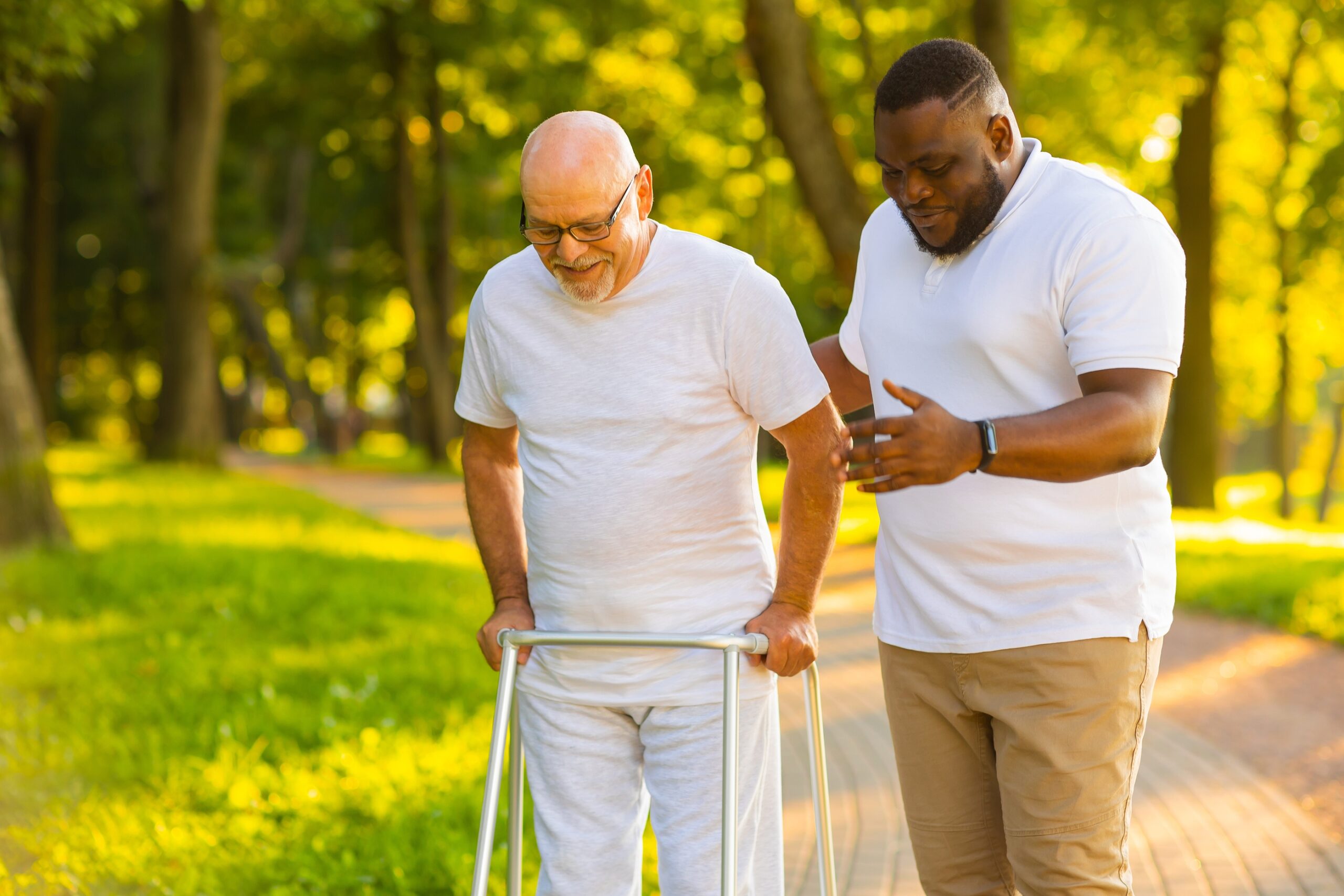 Elderly White male with walker being supported along a paved walkway by a Black male volunteer