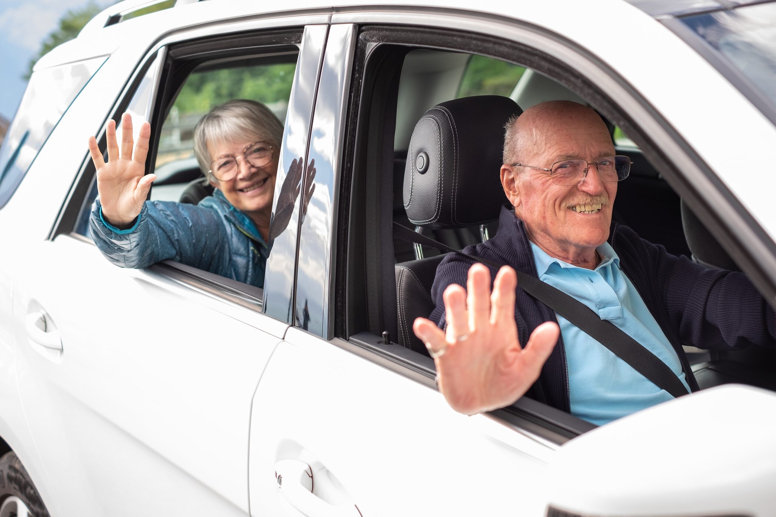 Happy senior woman and male volunteer driver smiling and waving out windows of their car